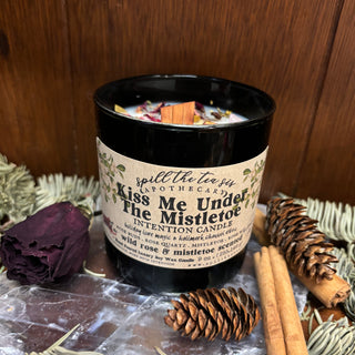 Candle in a decorative container with a label, surrounded by pine cones and cinnamon sticks on a wooden surface.